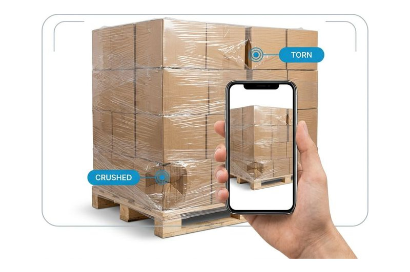 A warehouse worker photographing a pallet of cartons, with damage flagged on one carton